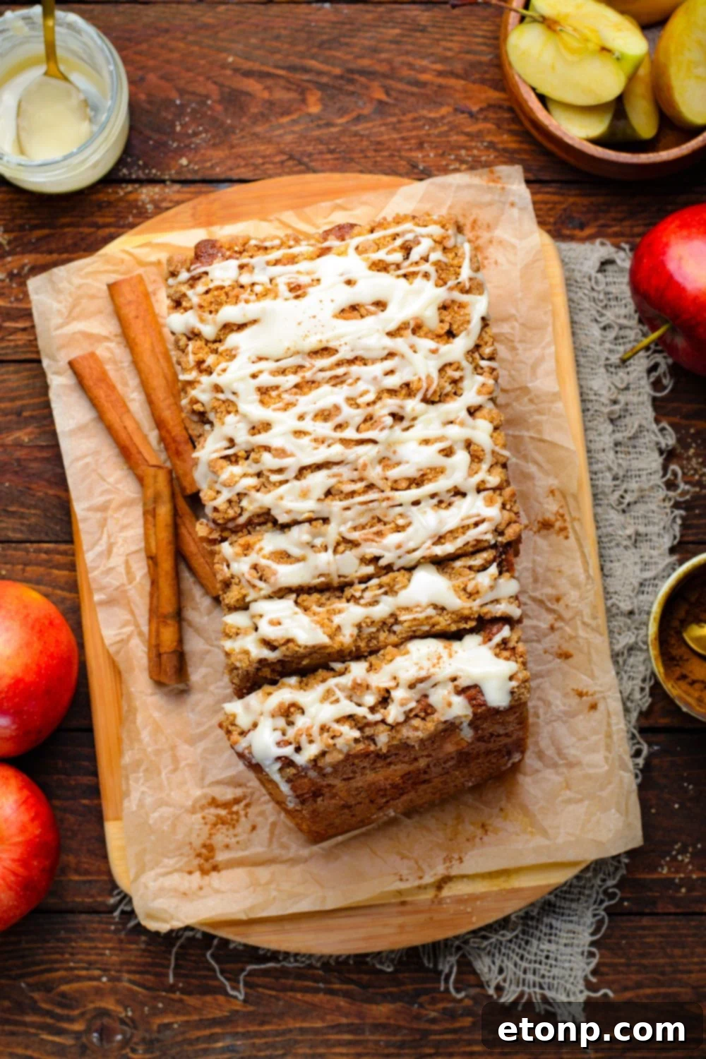 looking down on wooden table that has brown parchment paper in a rectangle with Apple Cinnamon Loaf Cake sliced and placed on it and apples placed around the table and cinnamon sticks