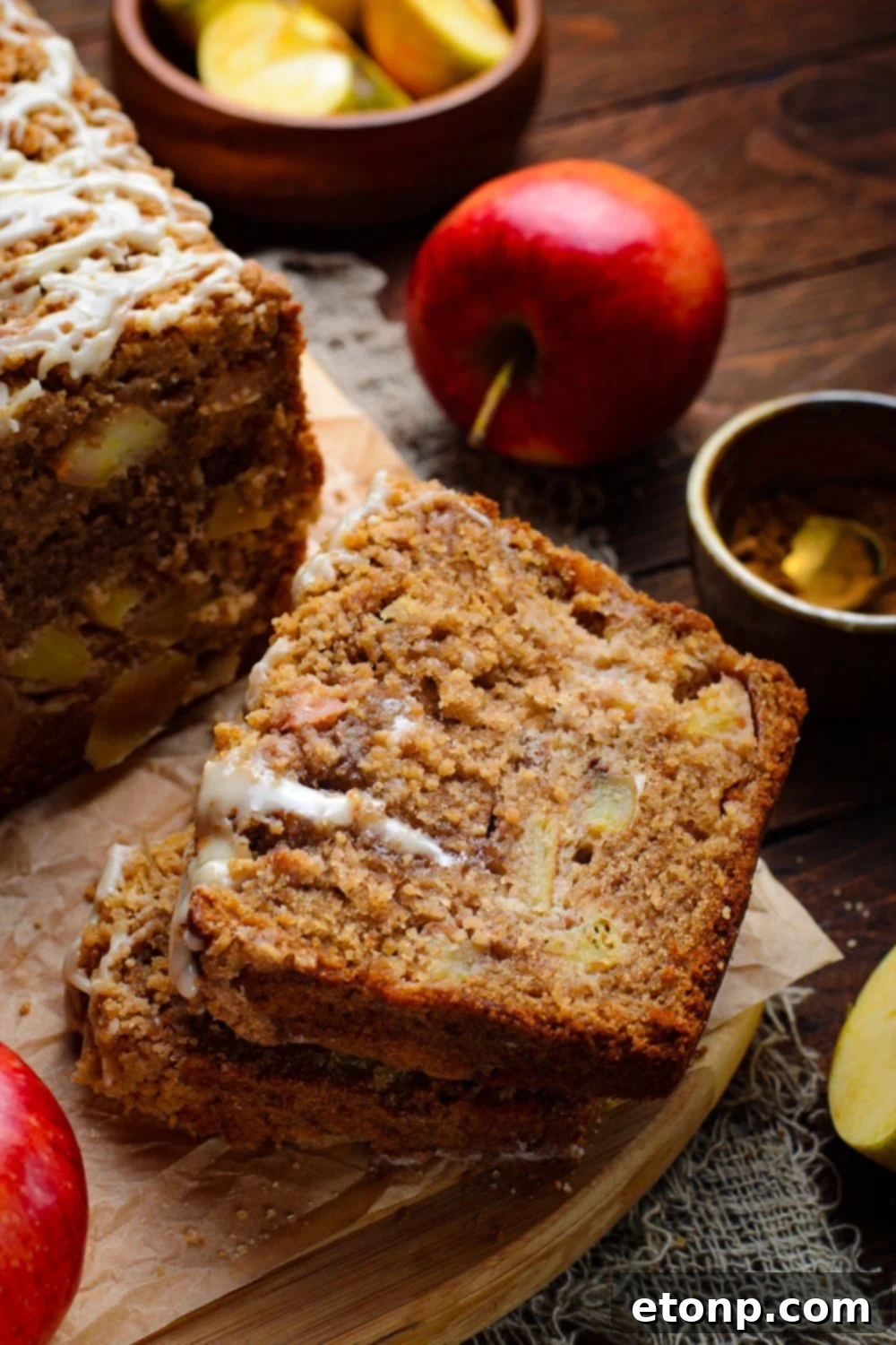 Apple Cinnamon Loaf Cake on a wooden table and some of the slices are stacked on top of one another with apples sliced in a wood bowl and a whole apple to side