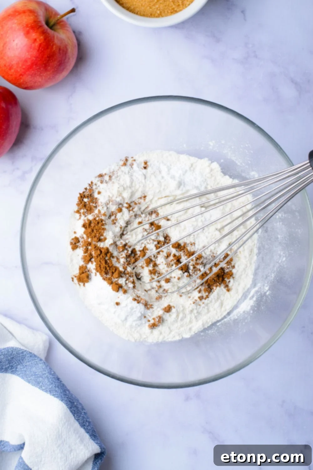 flour and cinnamon mixture in a glass bowl on marble counter with two apples set to the side on table
