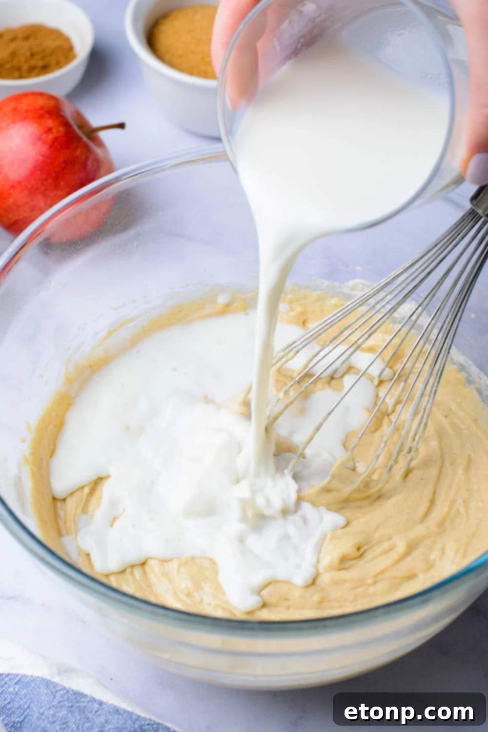 wet batter in a glass bowl with some milk pouring in and a whisk to the side. A red apple is on the table as well