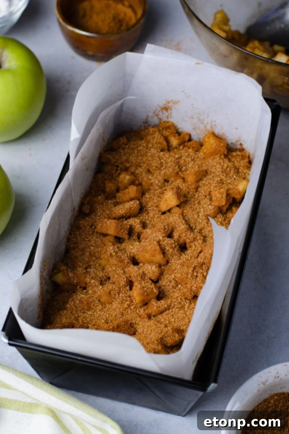cinnamon mixture and diced apples inside a loaf pan sitting on marble table with green apples to side of pan