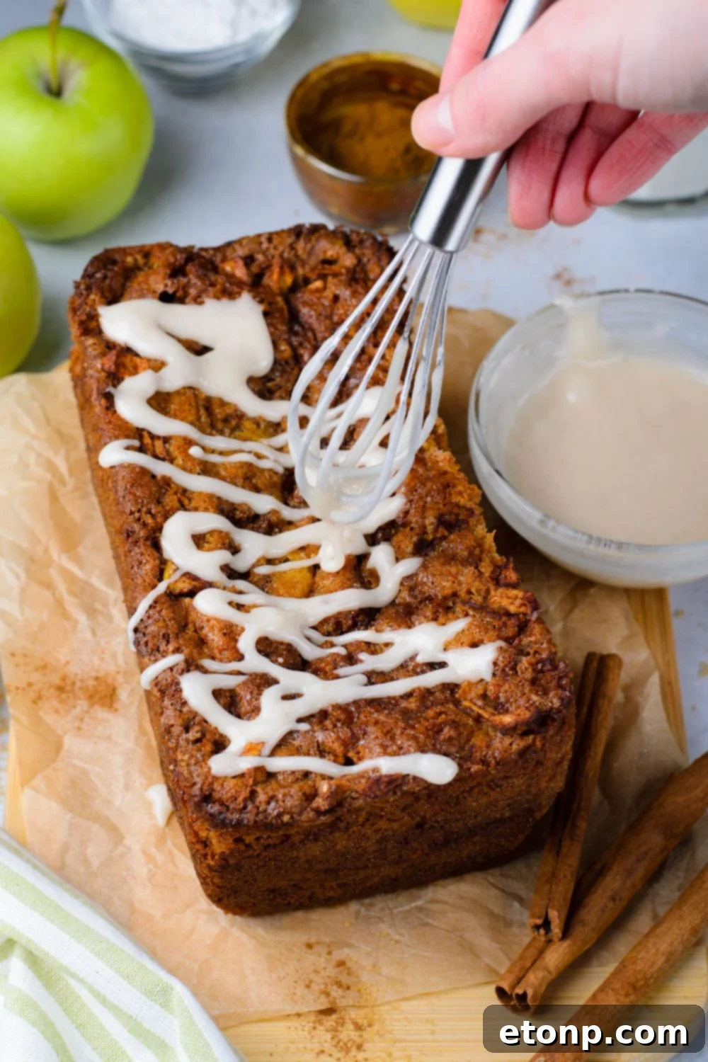 amish apple bread on table with a whisk drizzling glaze over the top with a small bowl of glaze to side