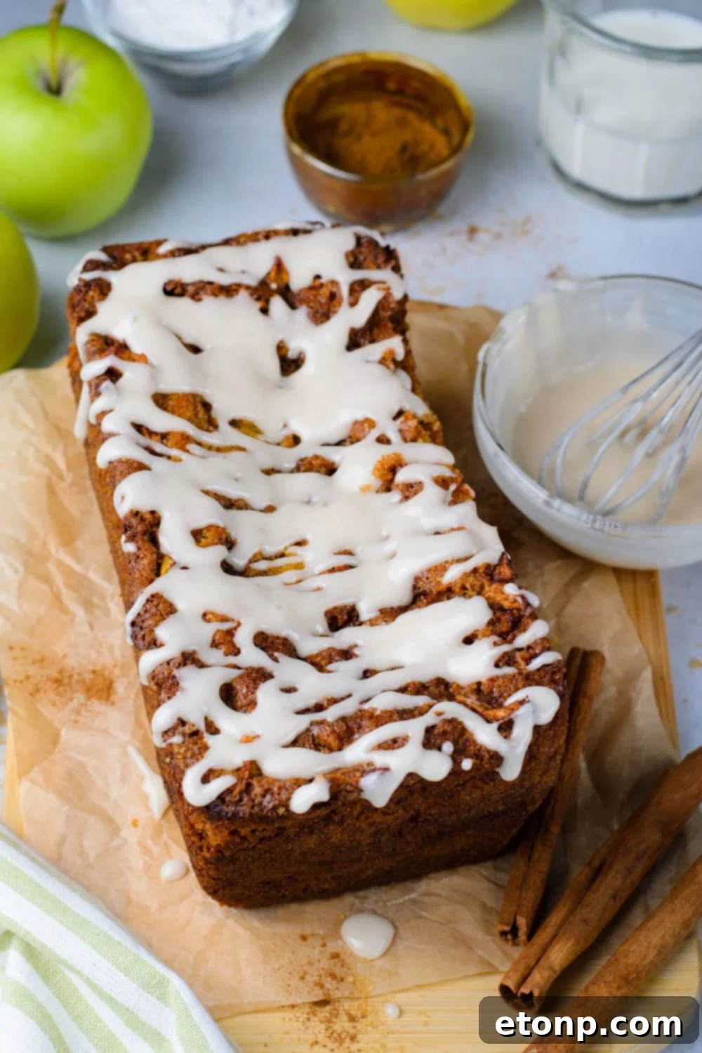 Amish Apple Fritter Bread on brown parchment paper on table with a bowl of glaze to side and cinnamon sticks in front of loaf