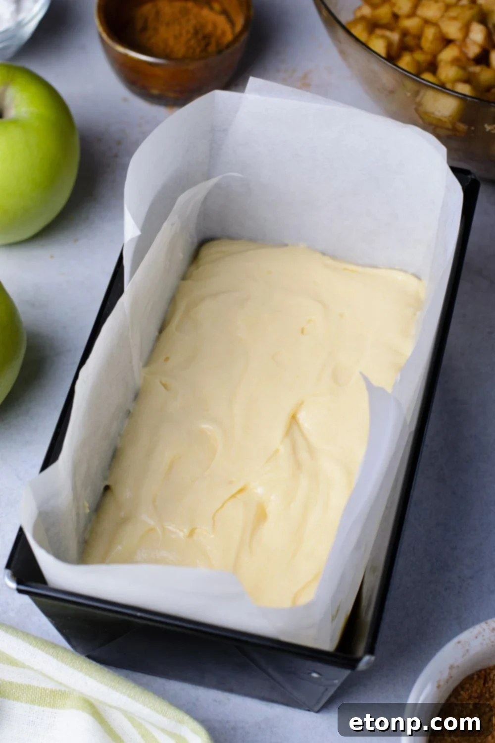loaf pan with parchment paper in it with a bit of bread batter inside. Apples, nuts, cinnamon and more around the pan to decorate the table