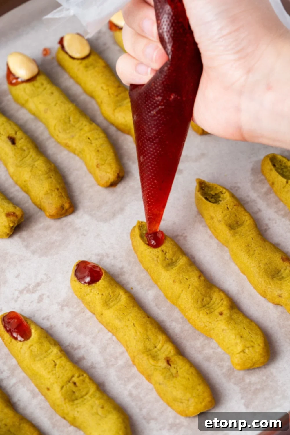 piping some jam on the tip of each of the Witch Finger Cookies on the baking tray 