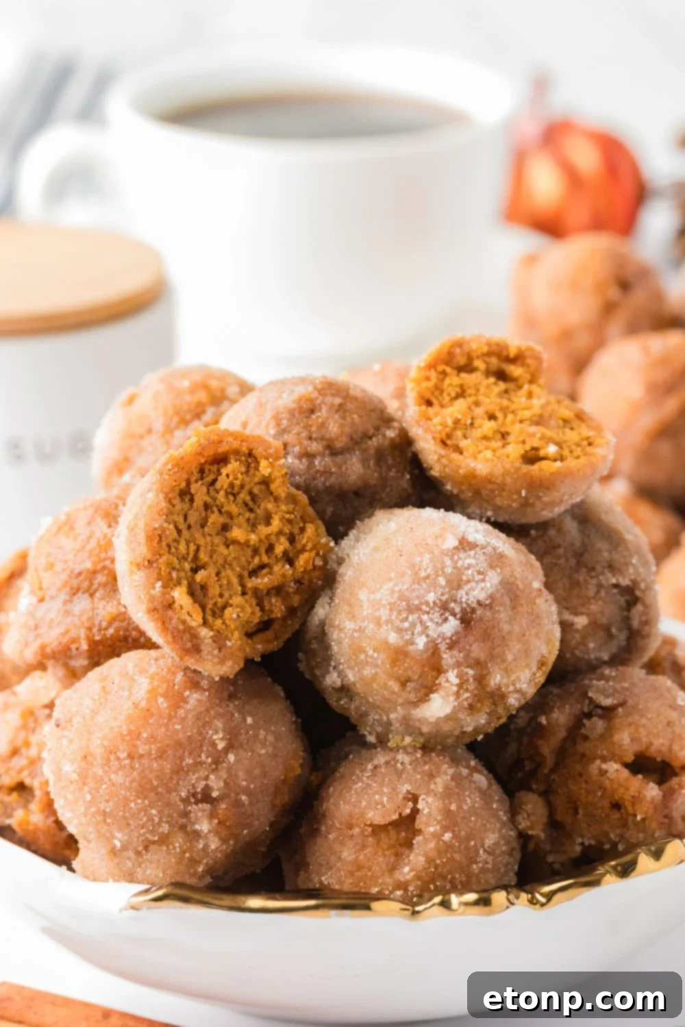 Pumpkin Spice Donut Holes stacked in a bowl on the counter with a few cut in half, and a cup of coffee in the background of the image 