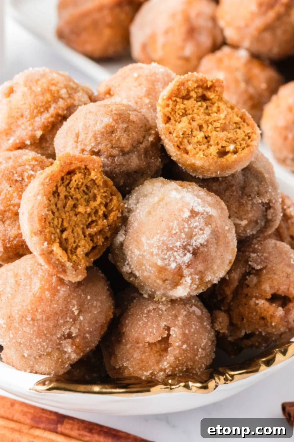 Pumpkin Spice Donut Holes stacked in a bowl on table with one cut in half, and a tray full of donuts behind it on table 