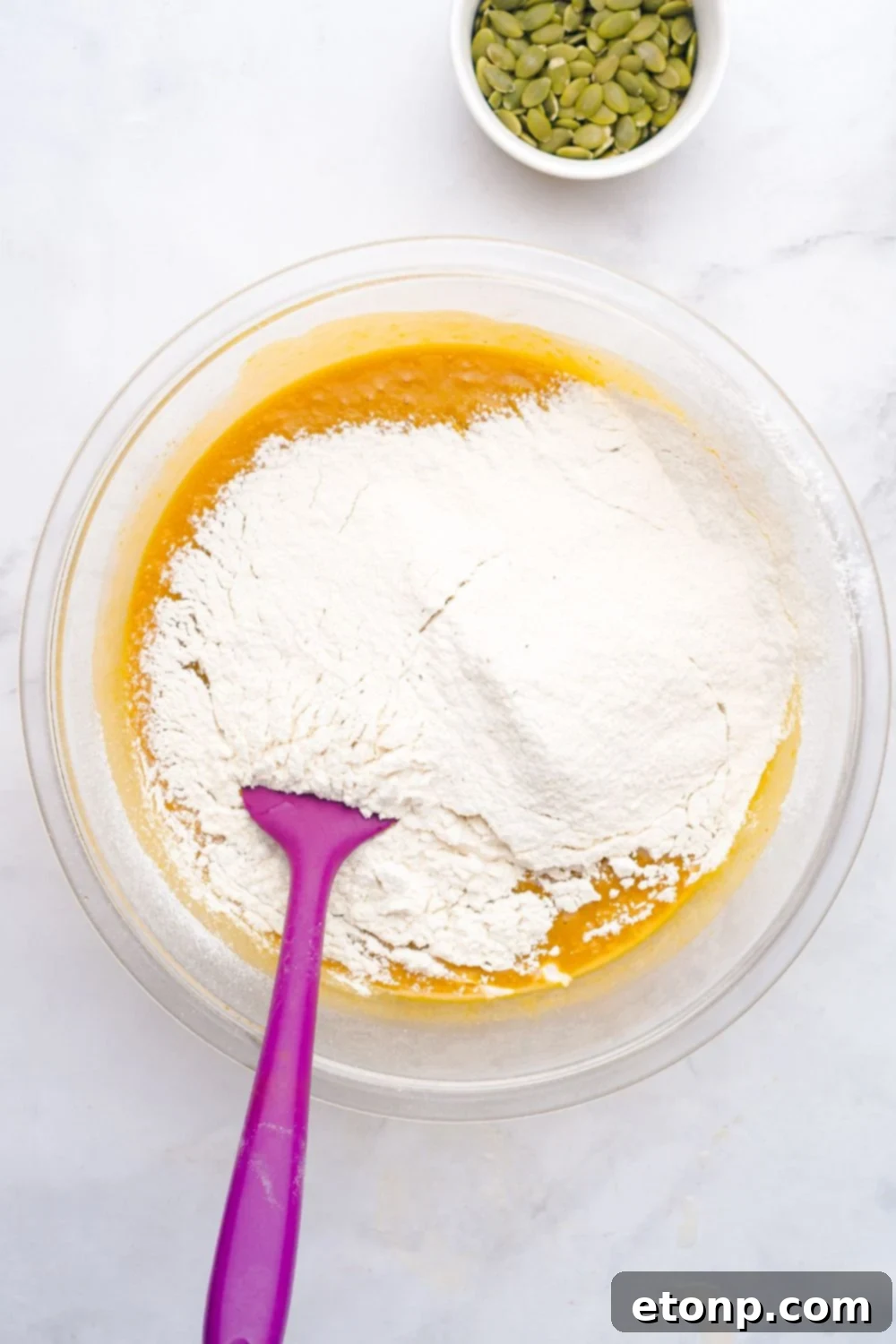 pumpkin bread batter in a glass bowl with flour on top and a purple spatula 