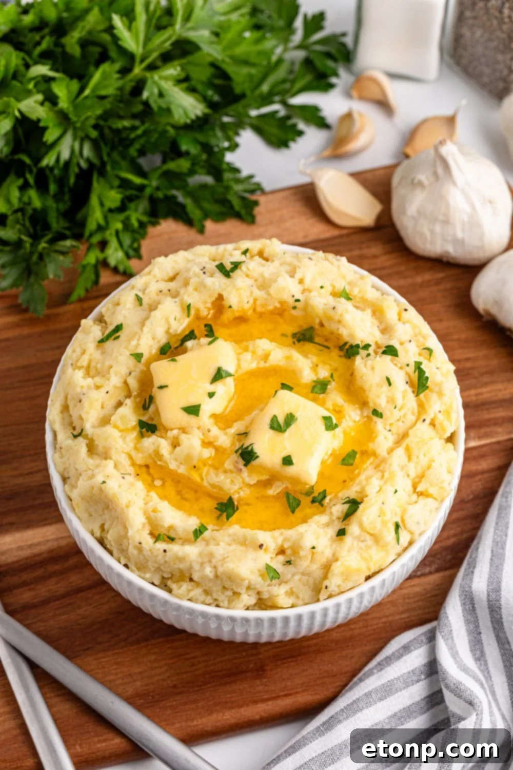 A wooden cutting board with a white bowl full of Creamy Garlic Mashed Potatoes, topped with melted butter and parsley. Fresh garlic and a kitchen towel are beside it