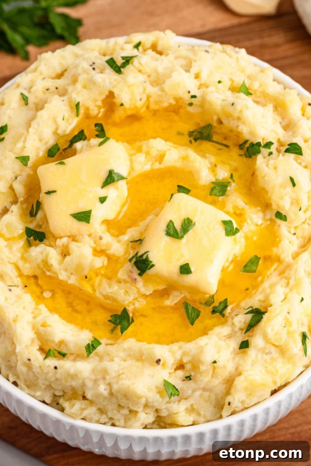 Close-up view of Creamy Garlic Mashed Potatoes in a white bowl on a wooden cutting board, topped with melted butter and fresh parsley