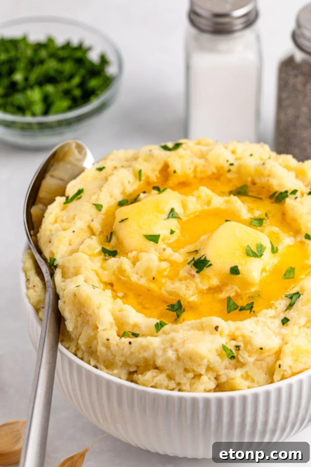 A white bowl on a table full of thick and creamy Garlic Mashed Potatoes with a spoon resting on its side, wedged into the bowl