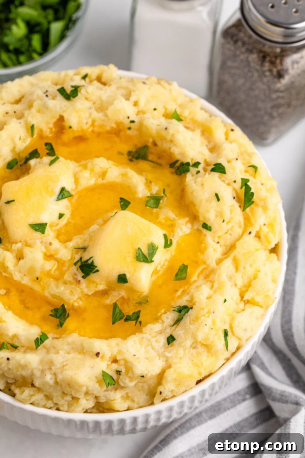 View of part of a white bowl on a table full of Creamy Garlic Mashed Potatoes with melted butter and chopped parsley on top. Salt and pepper shakers are to the side