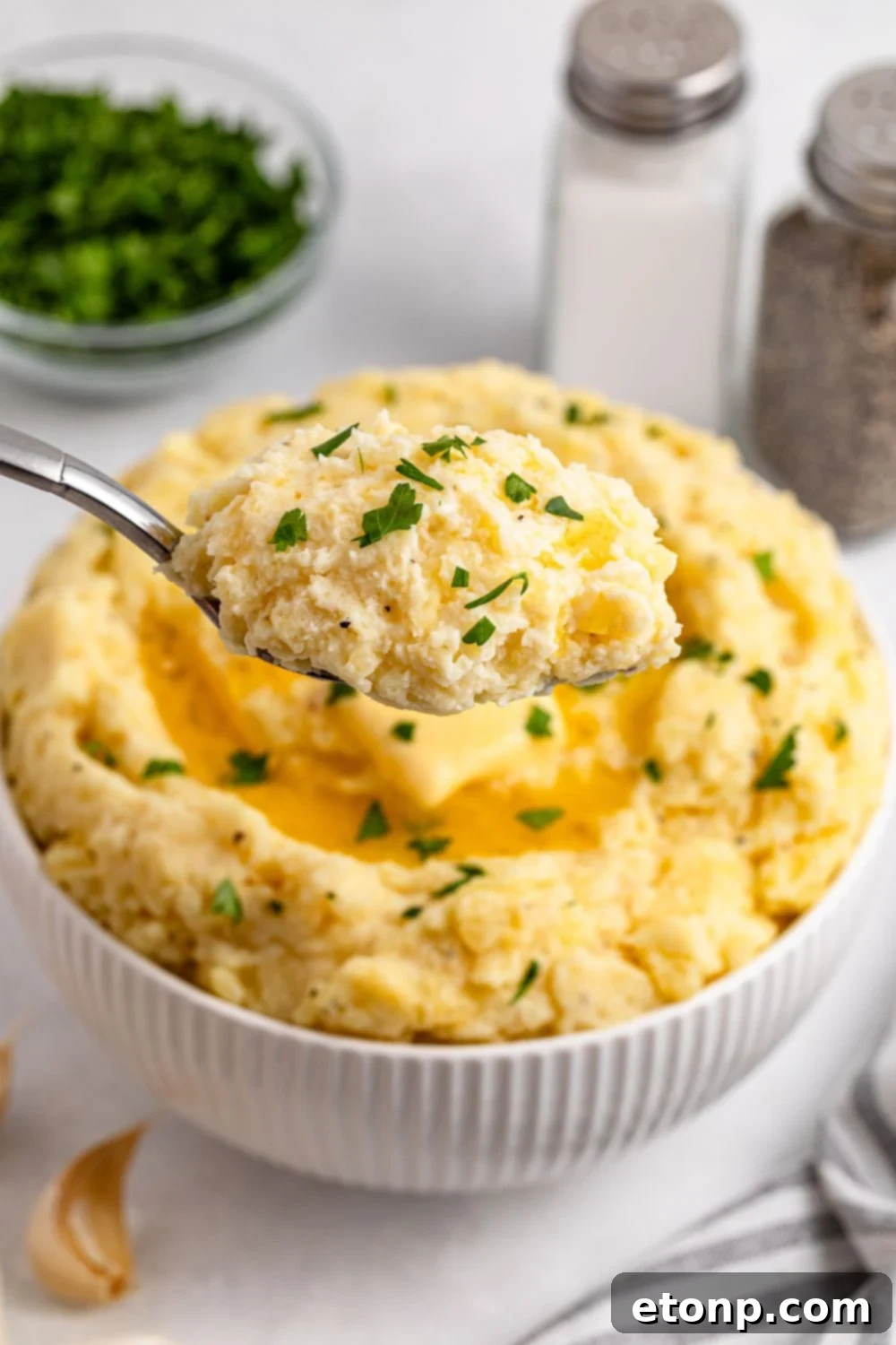 A spoon full of Creamy Garlic Mashed Potatoes with a bowl of more potatoes behind it on a white table. To the side is a salt and pepper shaker and a small glass bowl with chopped parsley