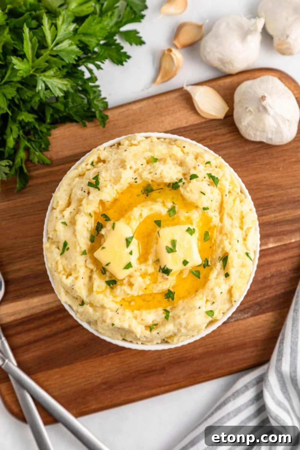 Looking down at a table to see a wooden cutting board with a bowl of Creamy Garlic Mashed Potatoes in the center. To the side are a bundle of parsley and fresh garlic cloves