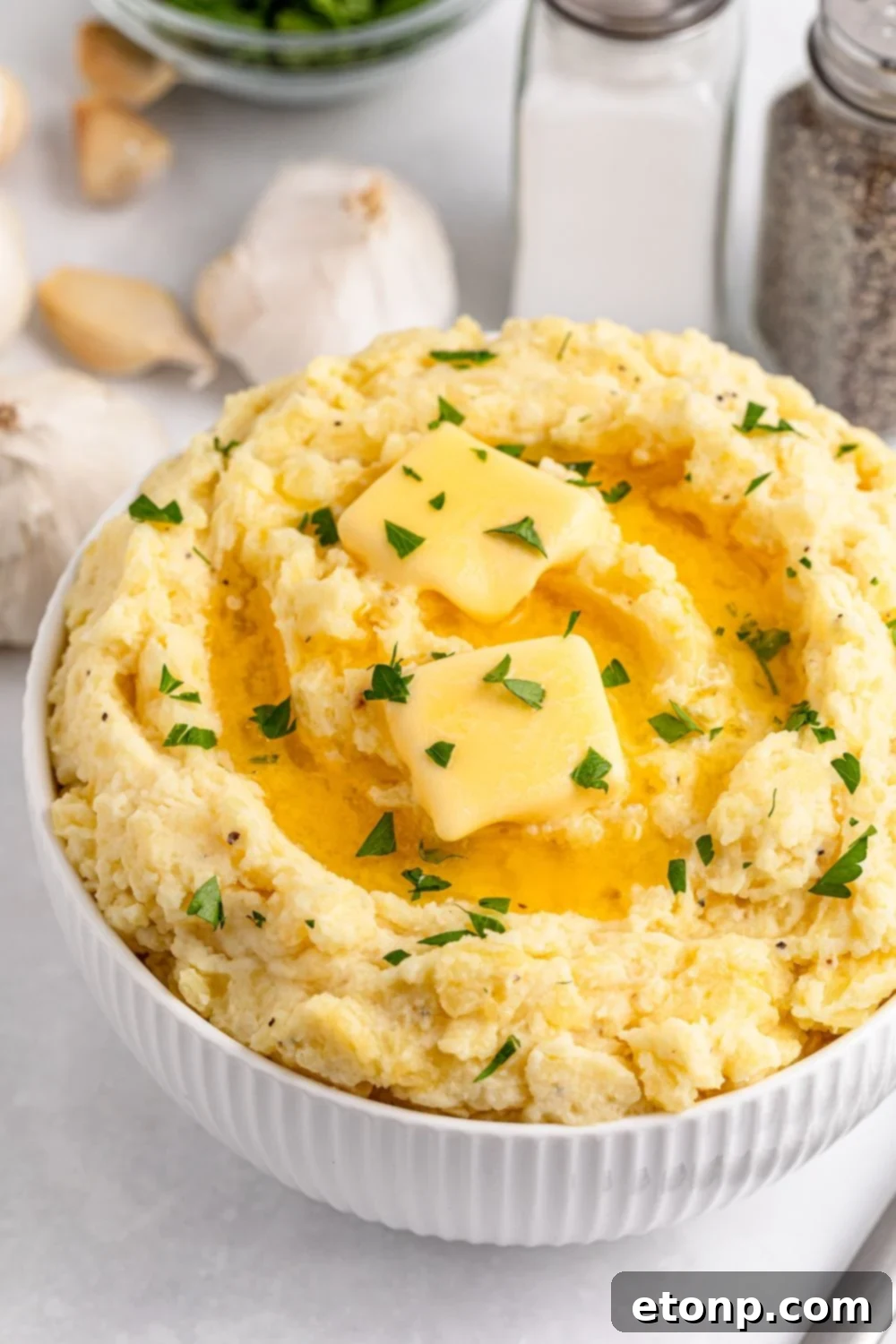 White table with salt and pepper shakers, a handful of garlic cloves, and a white bowl full of Creamy Garlic Mashed Potatoes