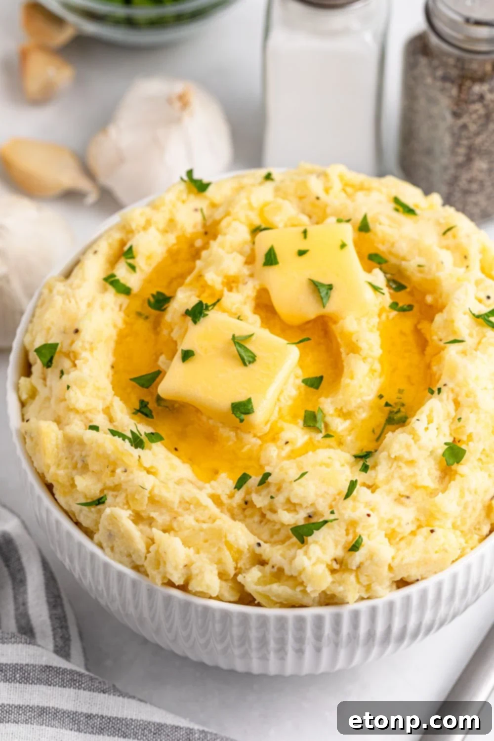 Creamy Garlic Mashed Potatoes in a bowl on a white table with fresh garlic and a striped towel to the side of the bowl