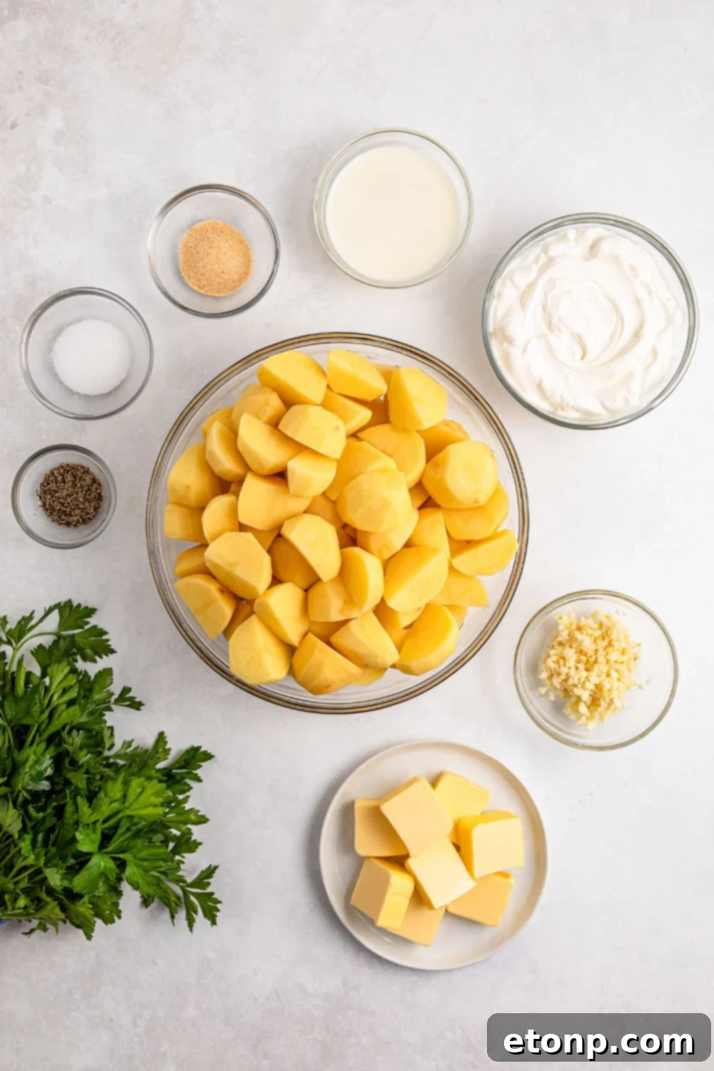 All the ingredients for Garlic Mashed Potatoes displayed on a white table in small bowls, including a bundle of parsley