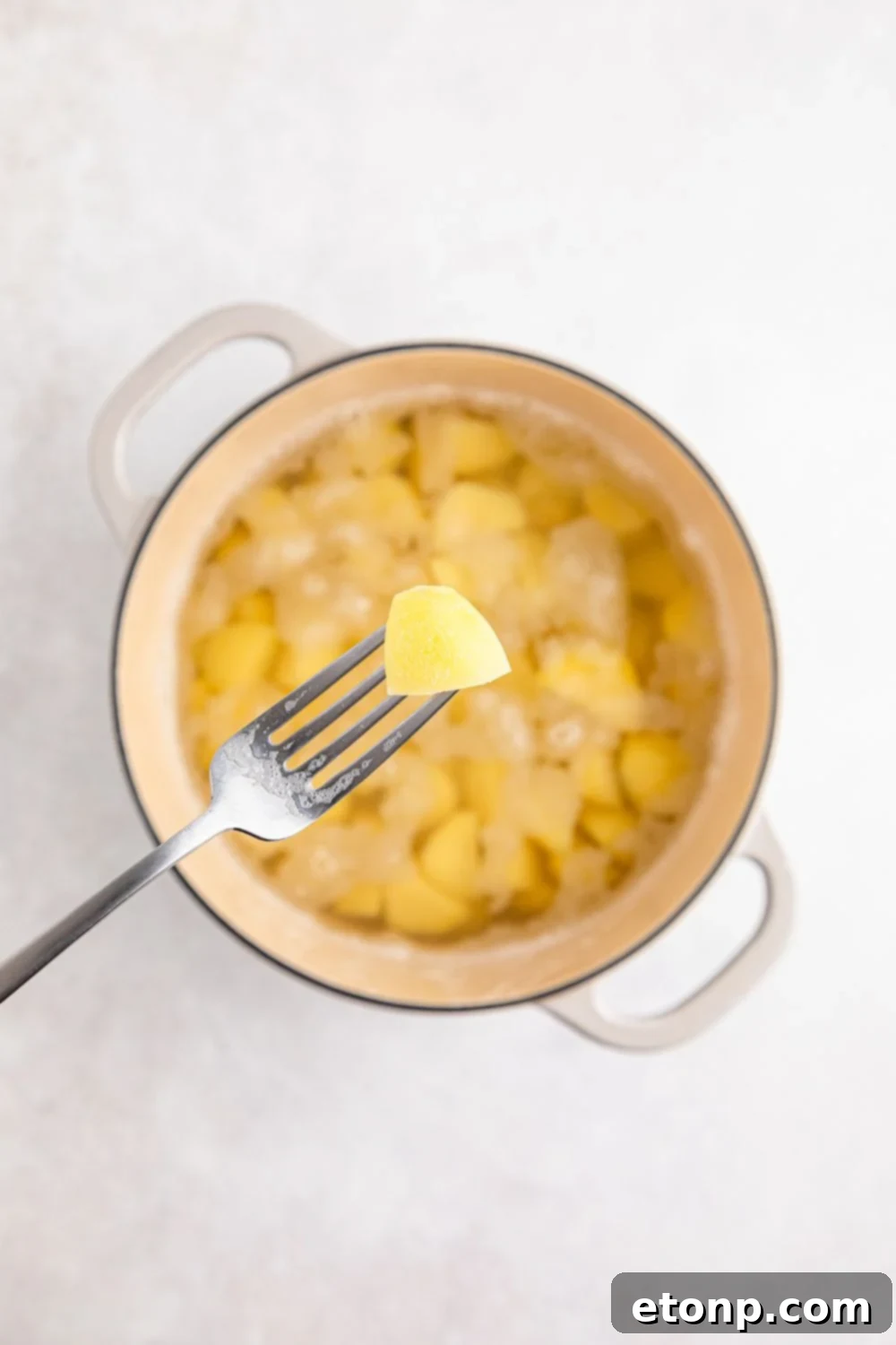 A fork holding a tender potato above a pot full of cooked potatoes and water, checking for doneness