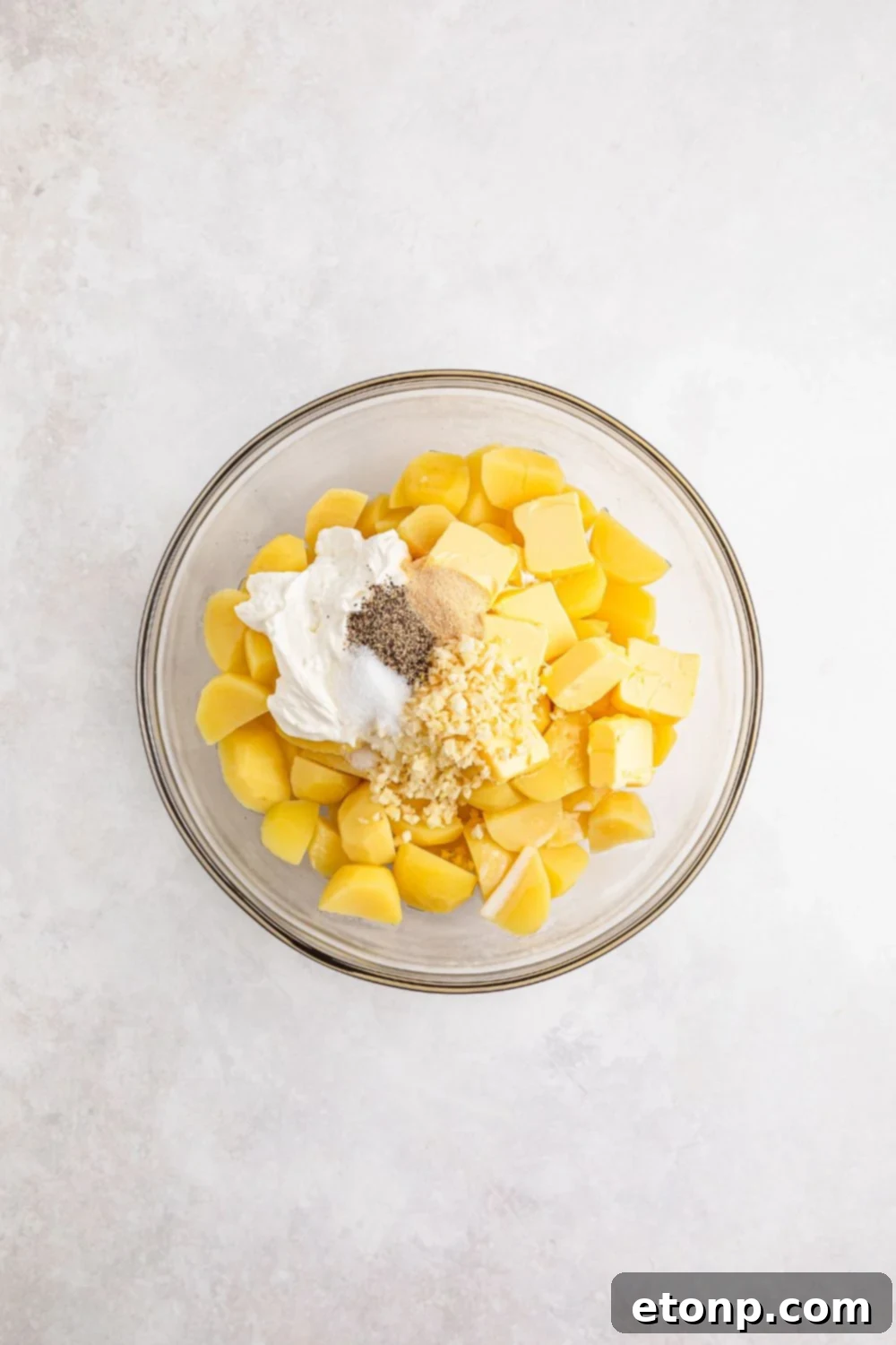 Cooked potatoes, sour cream, butter, minced garlic, and seasonings gathered in a glass bowl on a white table, ready for mashing