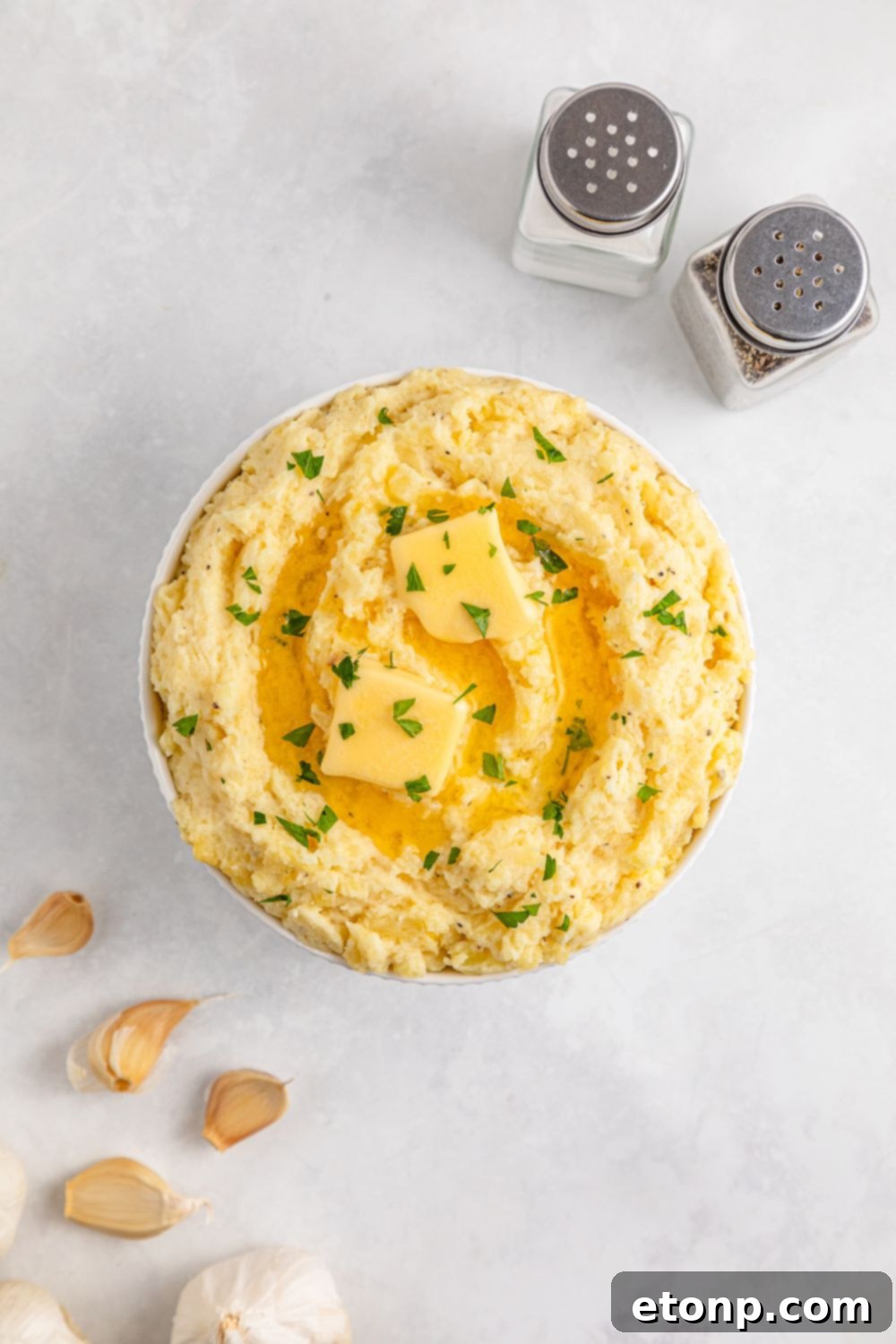 Large white bowl on table full of Creamy Garlic Mashed Potatoes, with fresh garlic cloves, salt, and pepper shakers to the side