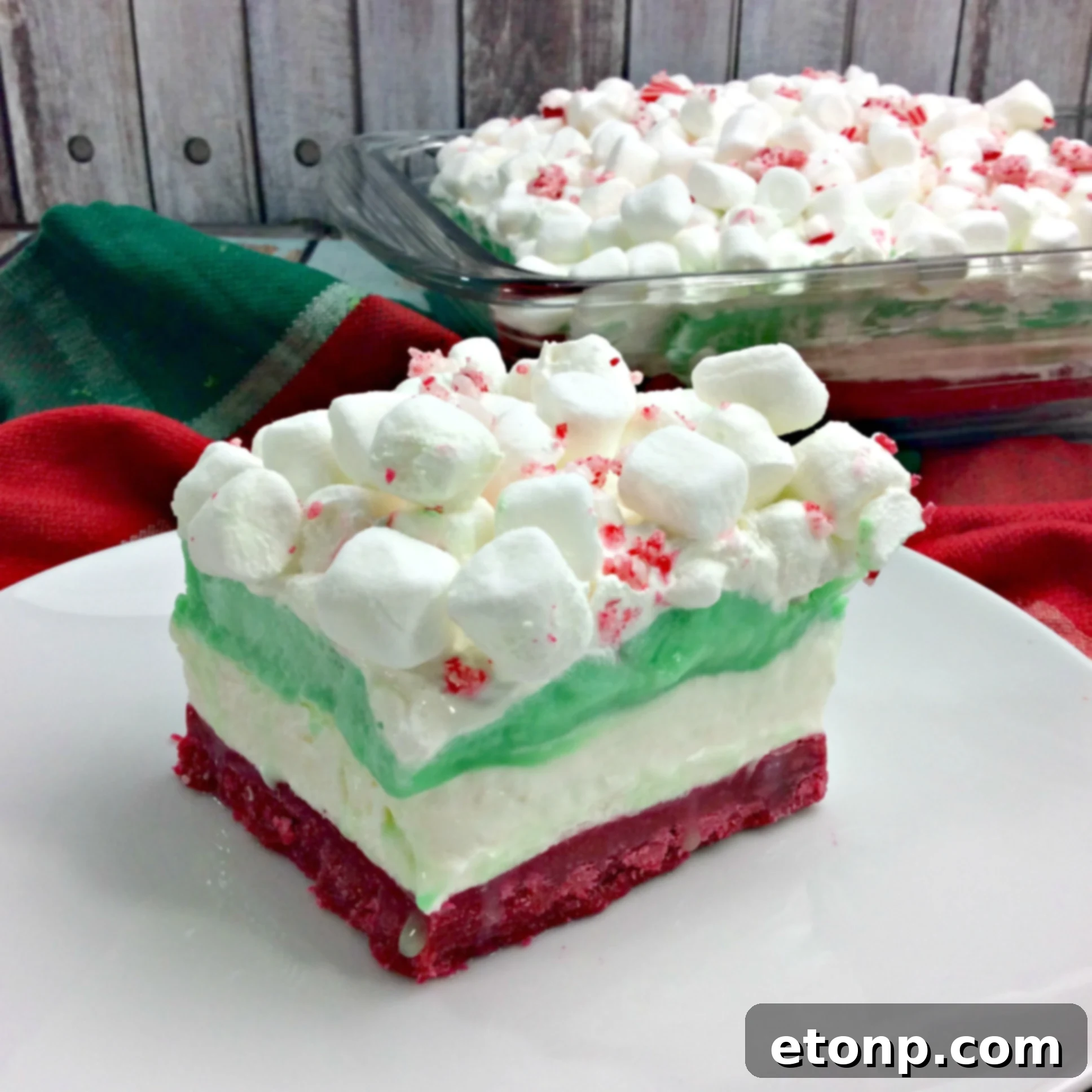 Side view of a generous serving of Christmas Cheesecake Lasagna on a plate, clearly showing the distinct colorful layers and garnished with candy cane pieces.
