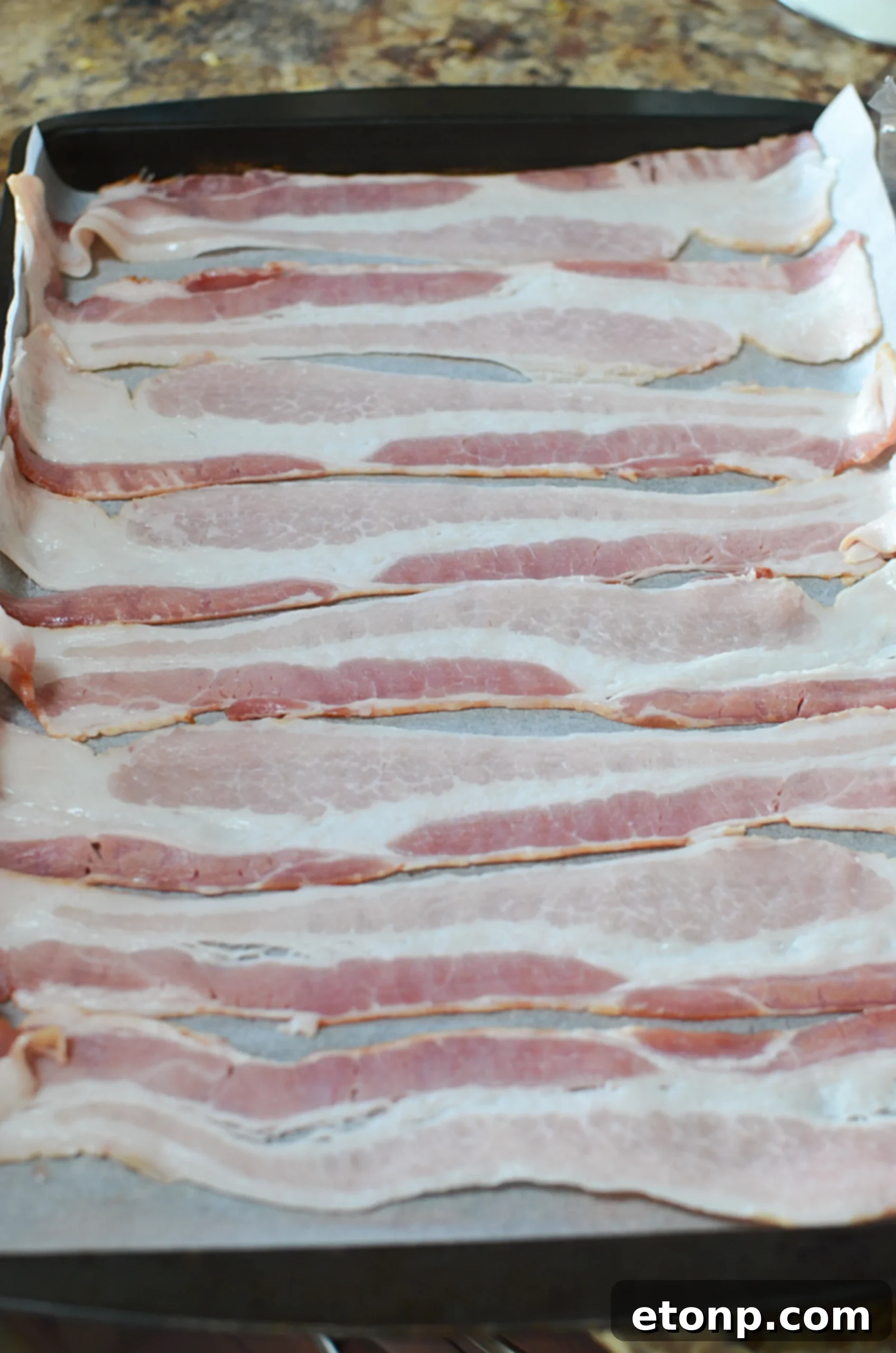 Raw bacon strips neatly laid out on parchment paper on a baking pan, preparing for oven baking