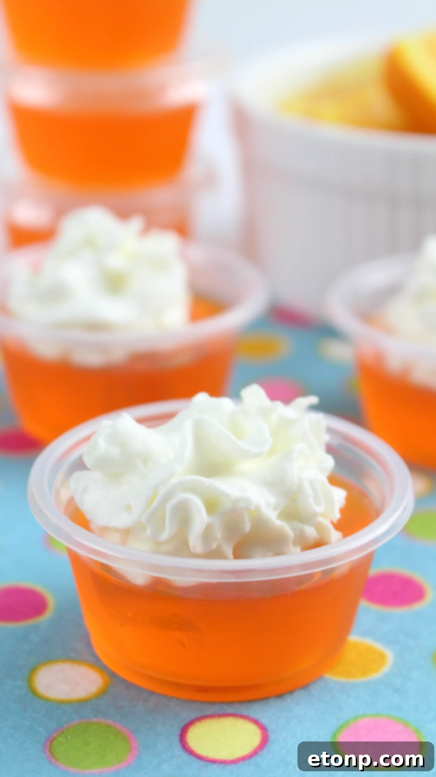 Close-up of vibrant orange creamsicle jello shots topped with whipped cream in clear plastic cups, set against a blurred party background.