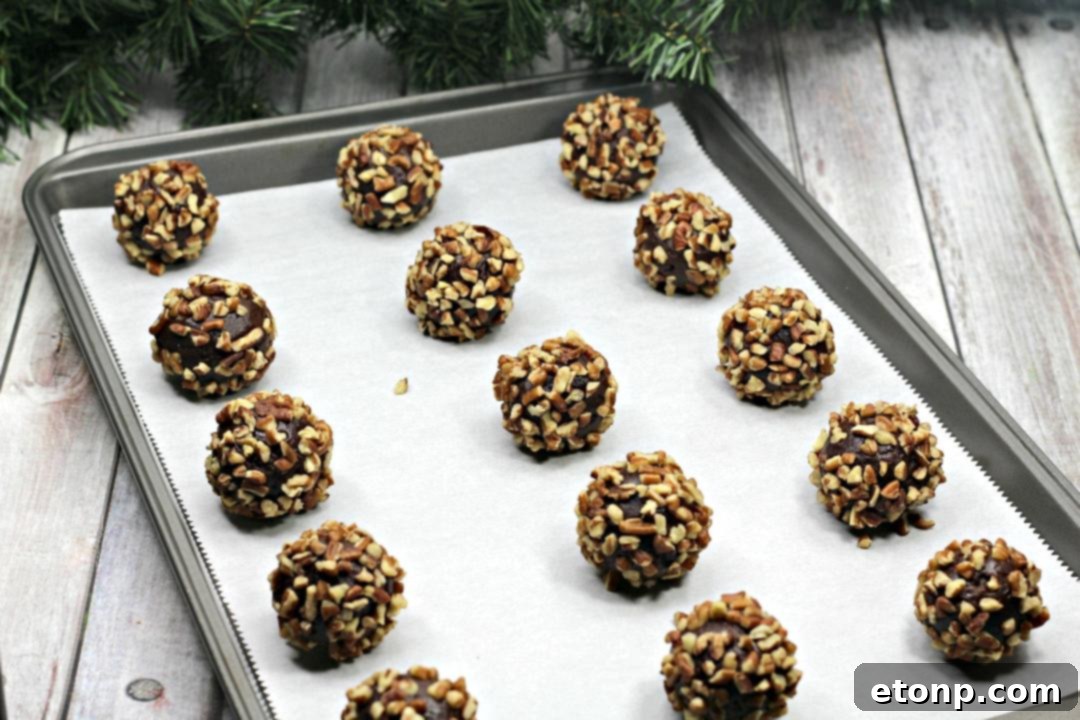 A hand gently pressing an indent into the center of a pecan-coated chocolate cookie dough ball on a baking sheet.