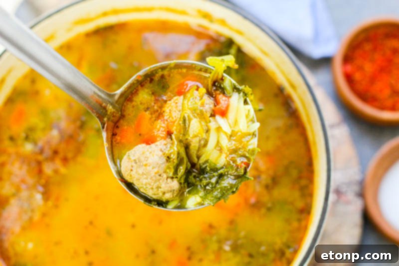 A close-up of a rustic wooden spoon stirring a pot of Tuscan meatball soup, highlighting the tender meatballs and fresh kale.