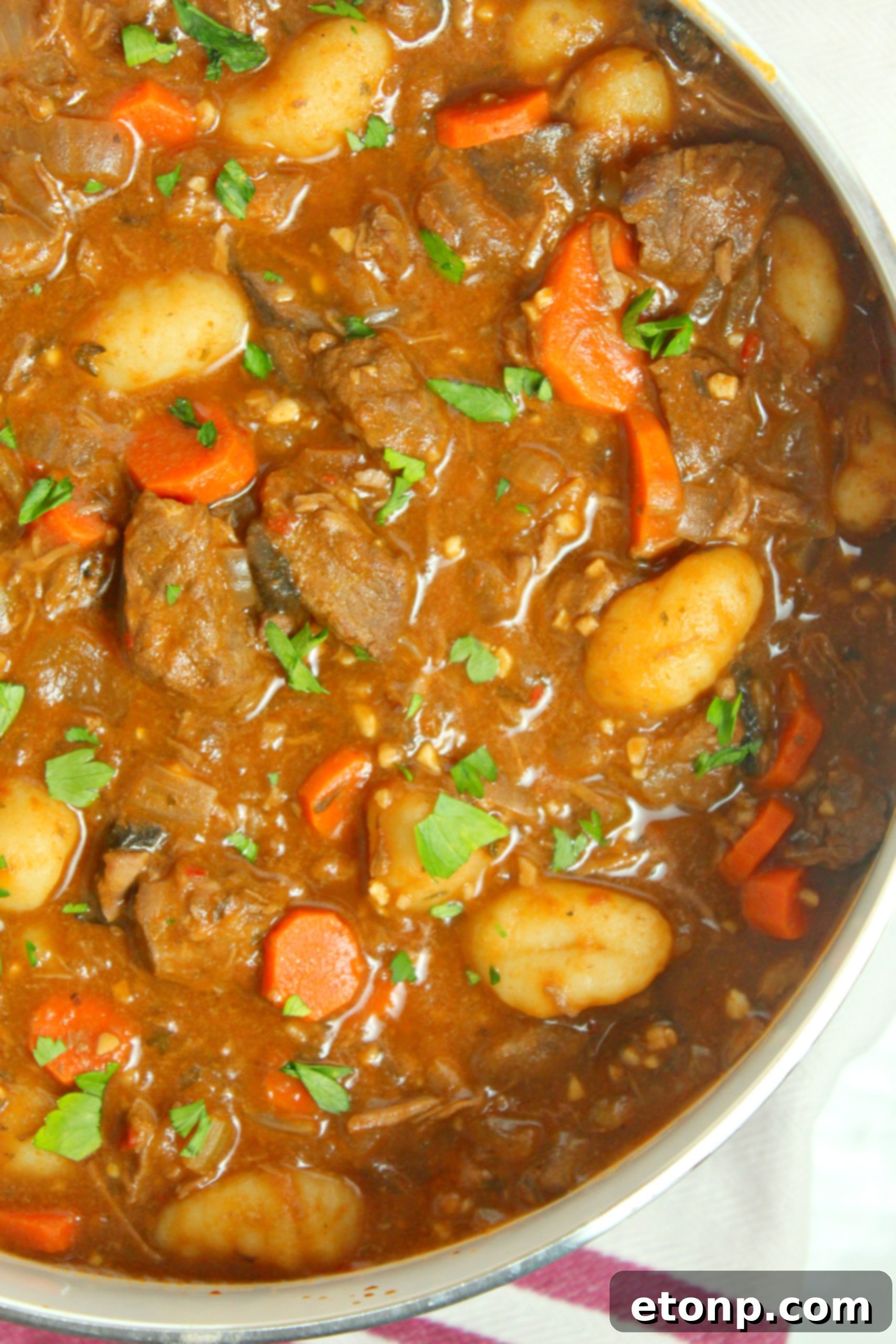 A steaming bowl of chunky beef and gnocchi soup with fresh parsley garnish, served on a rustic wooden table.
