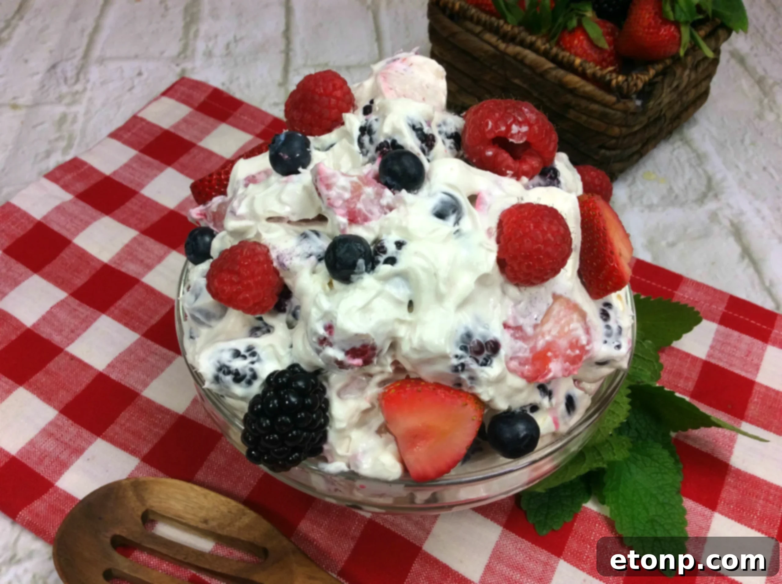 A rustic outdoor setting showing a large glass bowl of Cheesecake Summer Berry Salad on a wooden table, surrounded by other summer picnic items.