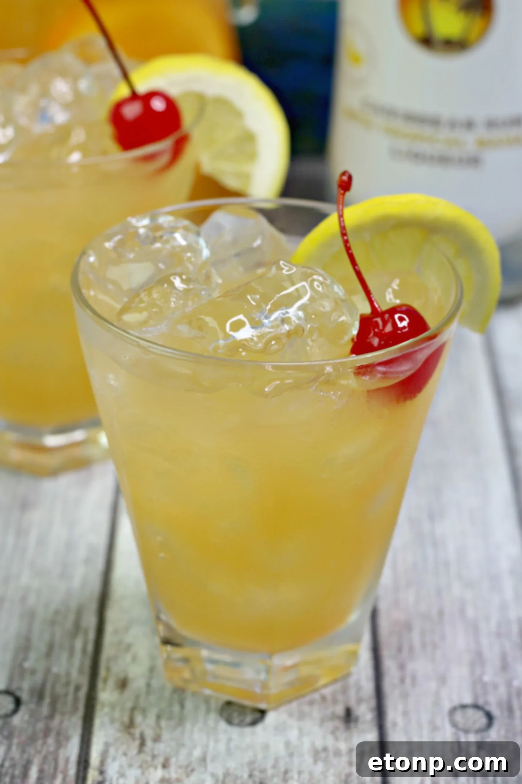 Two glasses of Banana Rum Punch, one clear and one slushy, garnished with fruit, sitting on a wooden table outdoors.