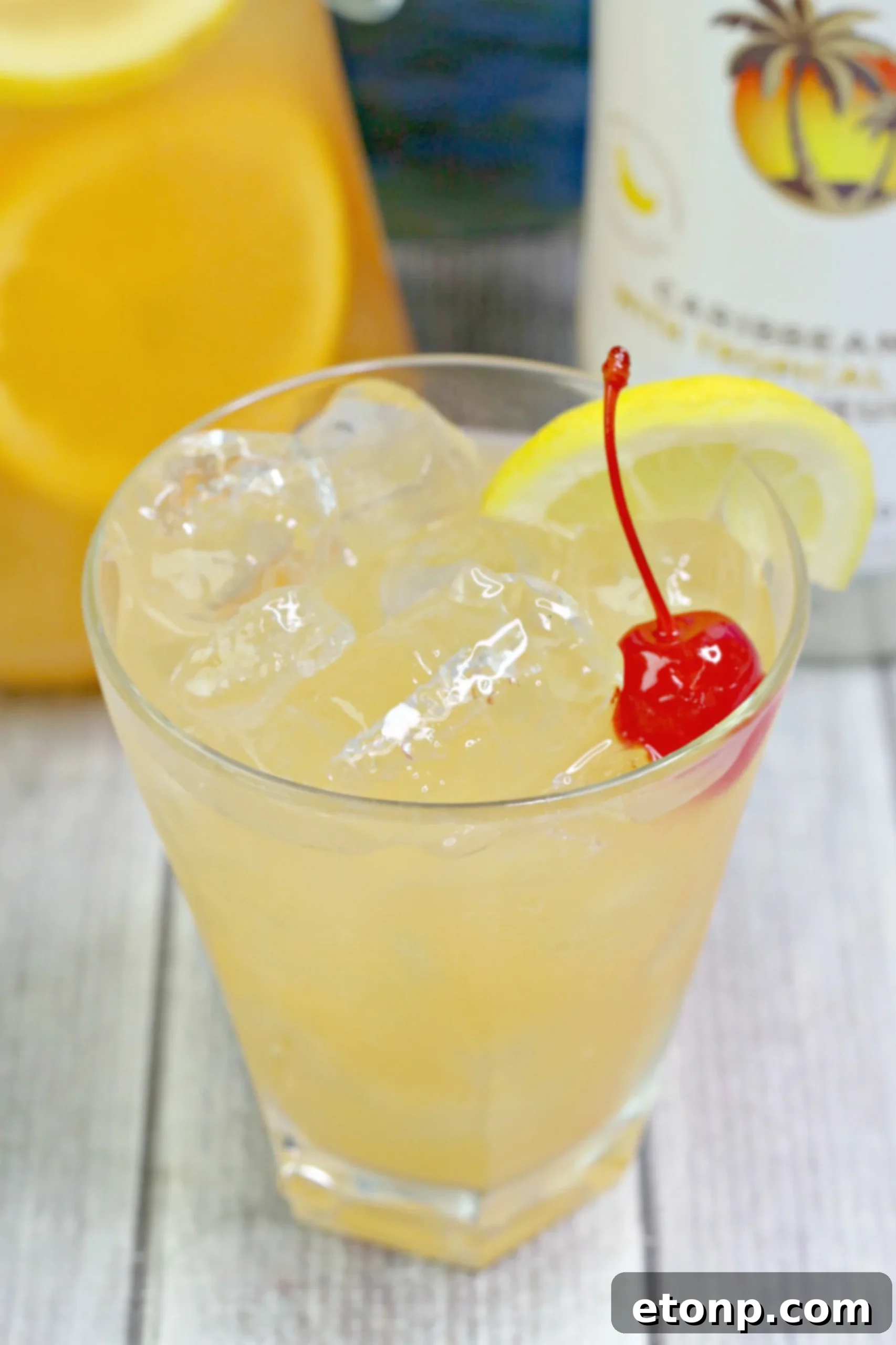 A pitcher of Banana Rum Punch being poured into a glass, with fresh fruit garnishes and ice.