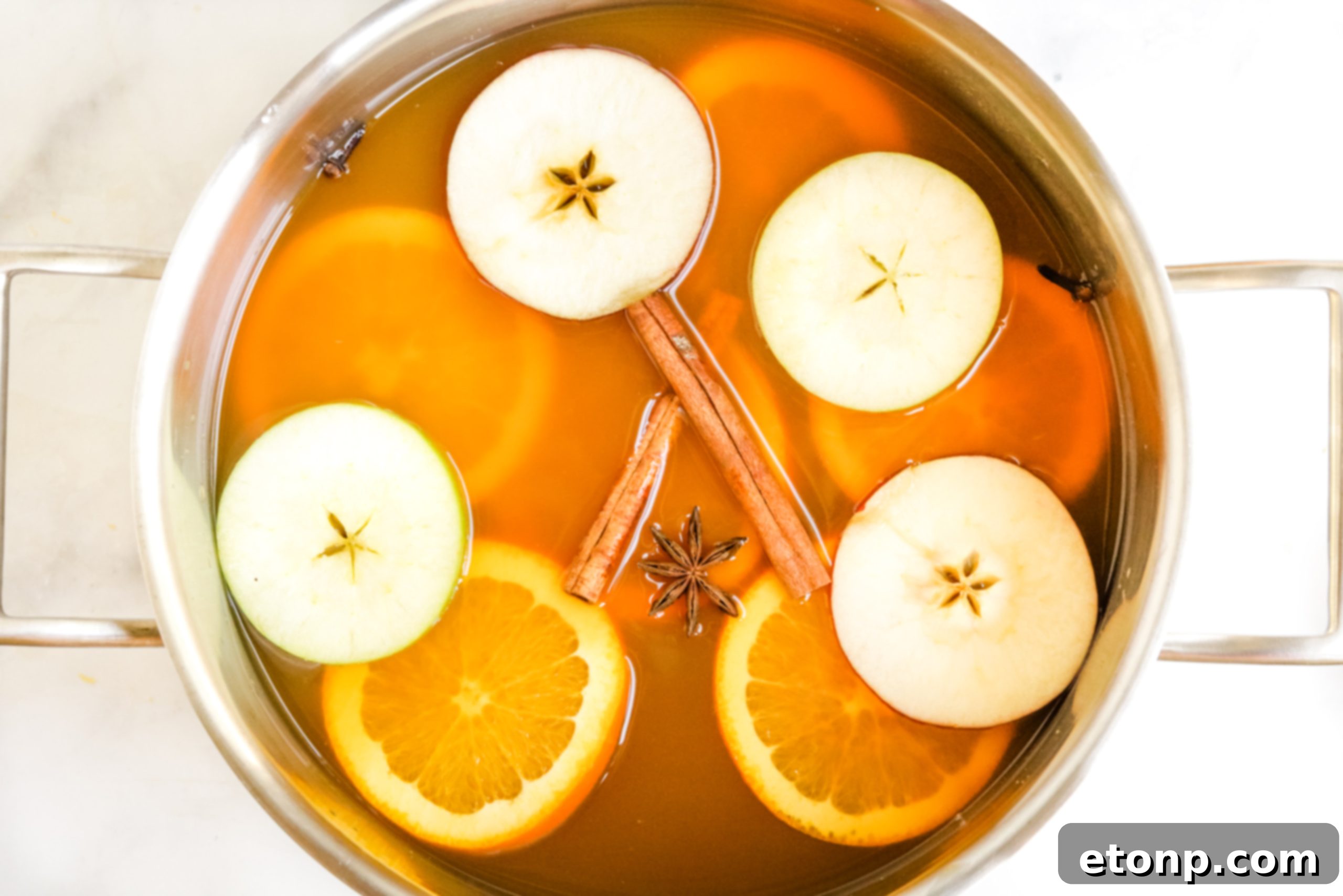 Close-up of apple and orange slices being added to a pot of mulled cider, alongside cinnamon sticks and whole cloves.