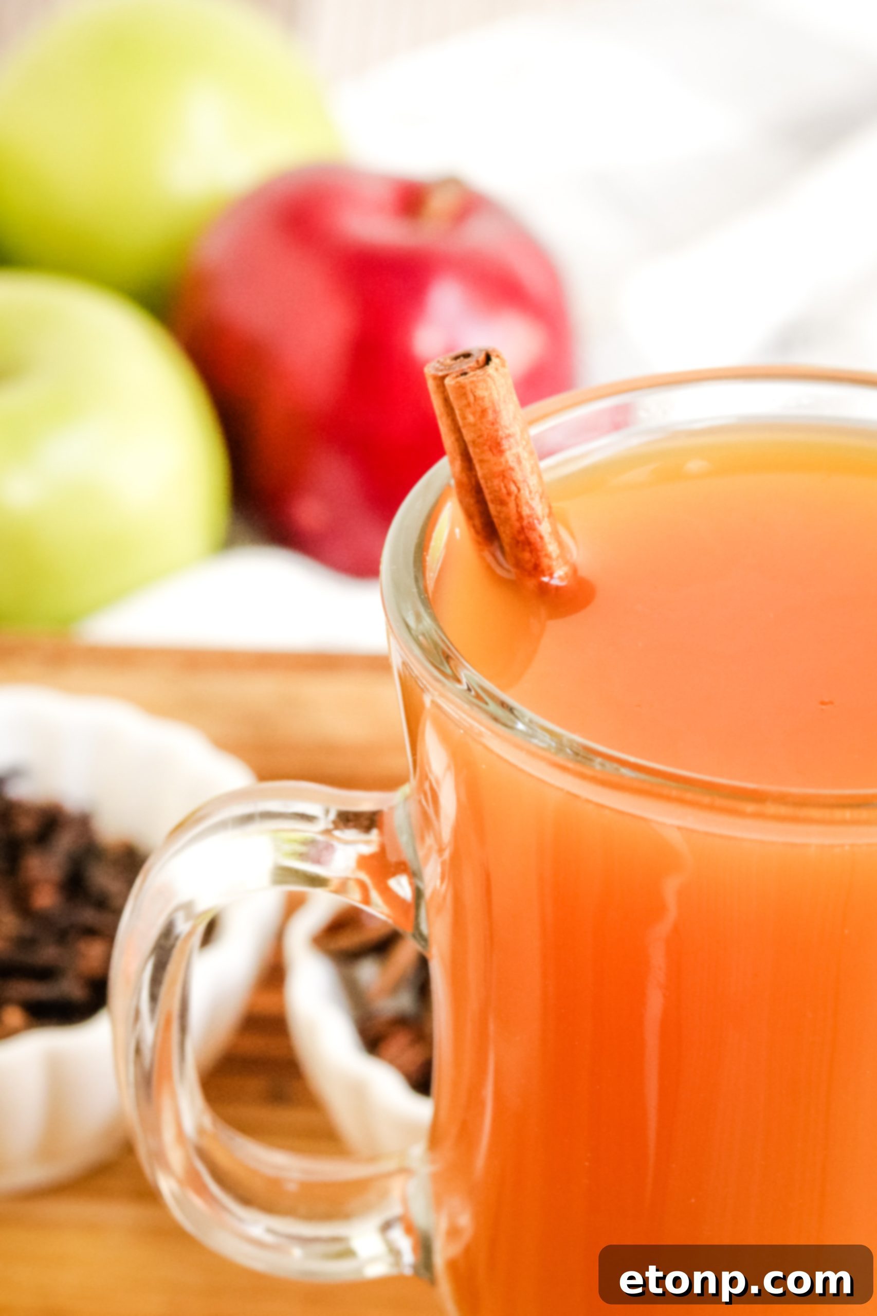 Two hands pouring hot mulled cider from a saucepan into a mug, with sliced fruit and spices visible.