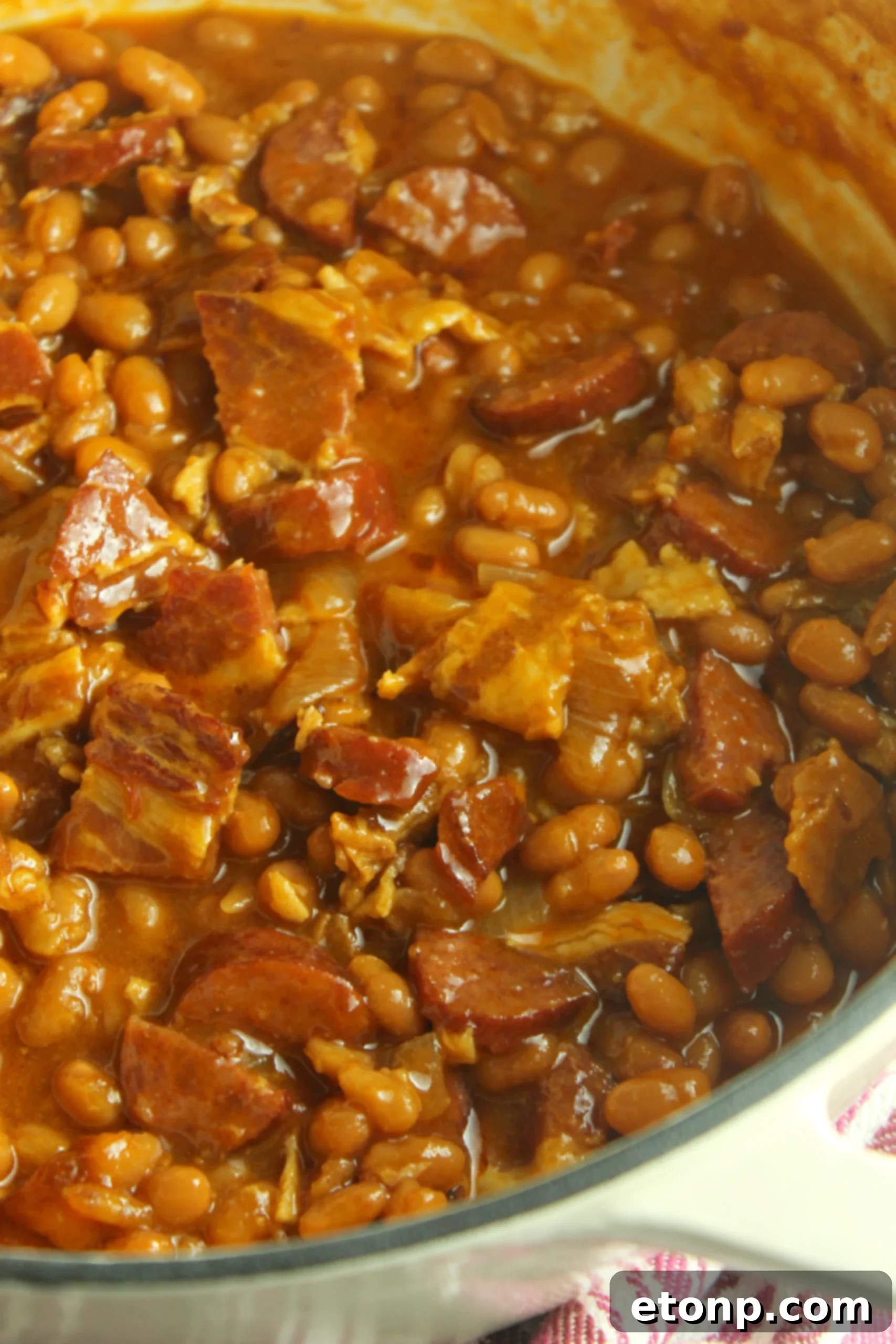 The ultimate baked beans with smoked sausage are pictured in a serving bowl, showcasing a rich, thick texture and hearty ingredients.