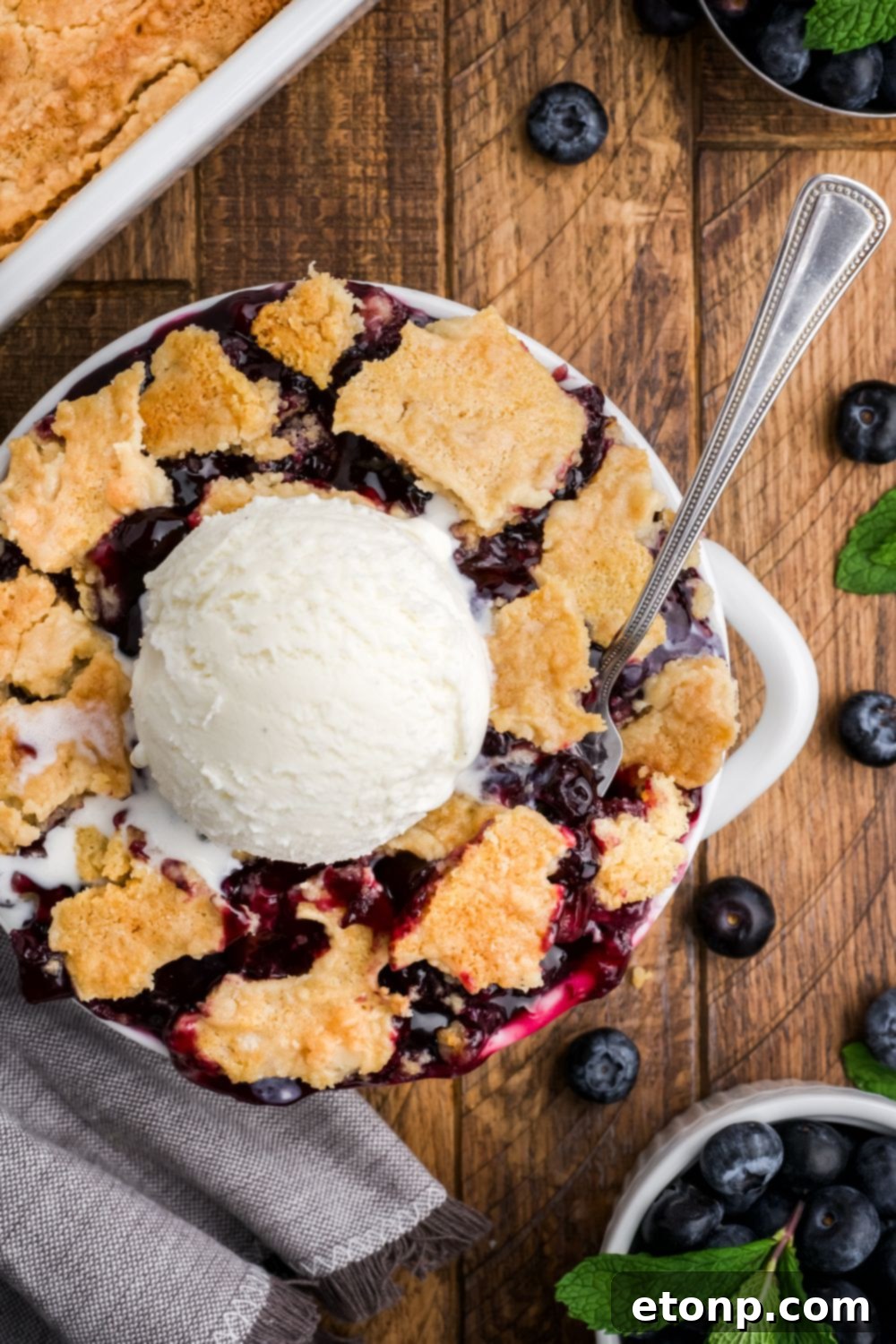 looking down on wooden table with a bowl of Blueberry Dump Cake with a scoop of ice cream in center