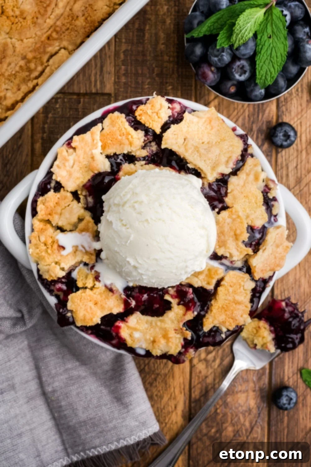 looking down on wooden table with a bowl full of dump blueberry cake with ice cream in the center and a bowl of blueberries to side