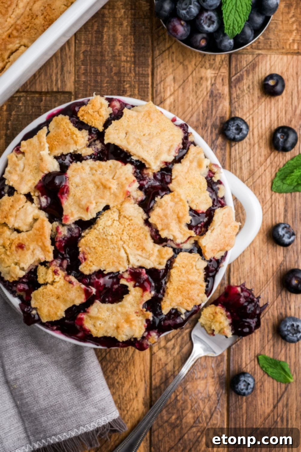 white bowl overflowing with blueberry dump cake and a fork with more cake with more blueberries scattered on table
