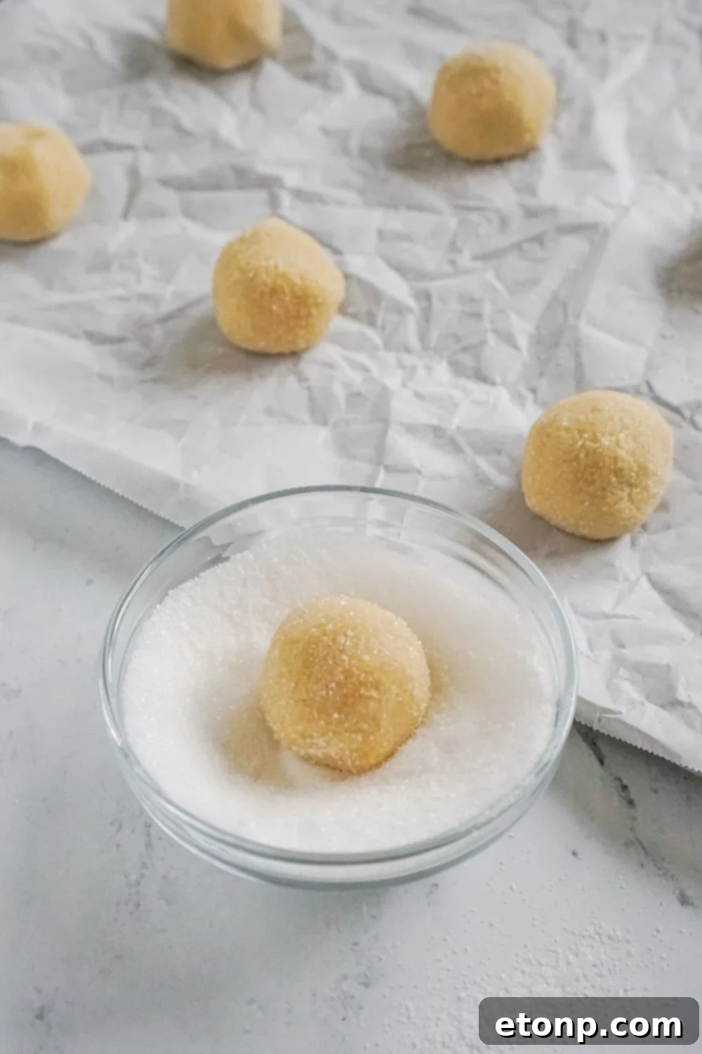 Cookie dough being rolled in a small bowl of sugar near a baking sheet.
