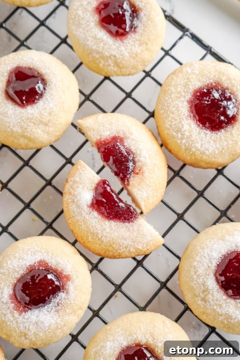 A close-up view of Raspberry Thumbprint Cookies on a cooling rack, with one cookie cut in half.