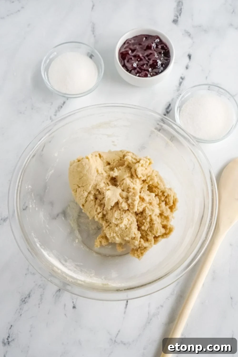 Cookie dough in a glass bowl, surrounded by other ingredient bowls on a marble countertop.