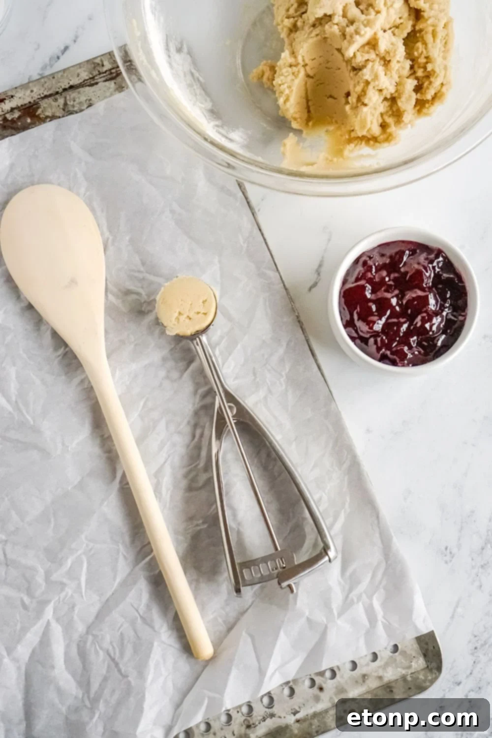 Raspberry Thumbprint Cookie dough in a glass bowl, with jam in a small bowl and a baking sheet nearby.