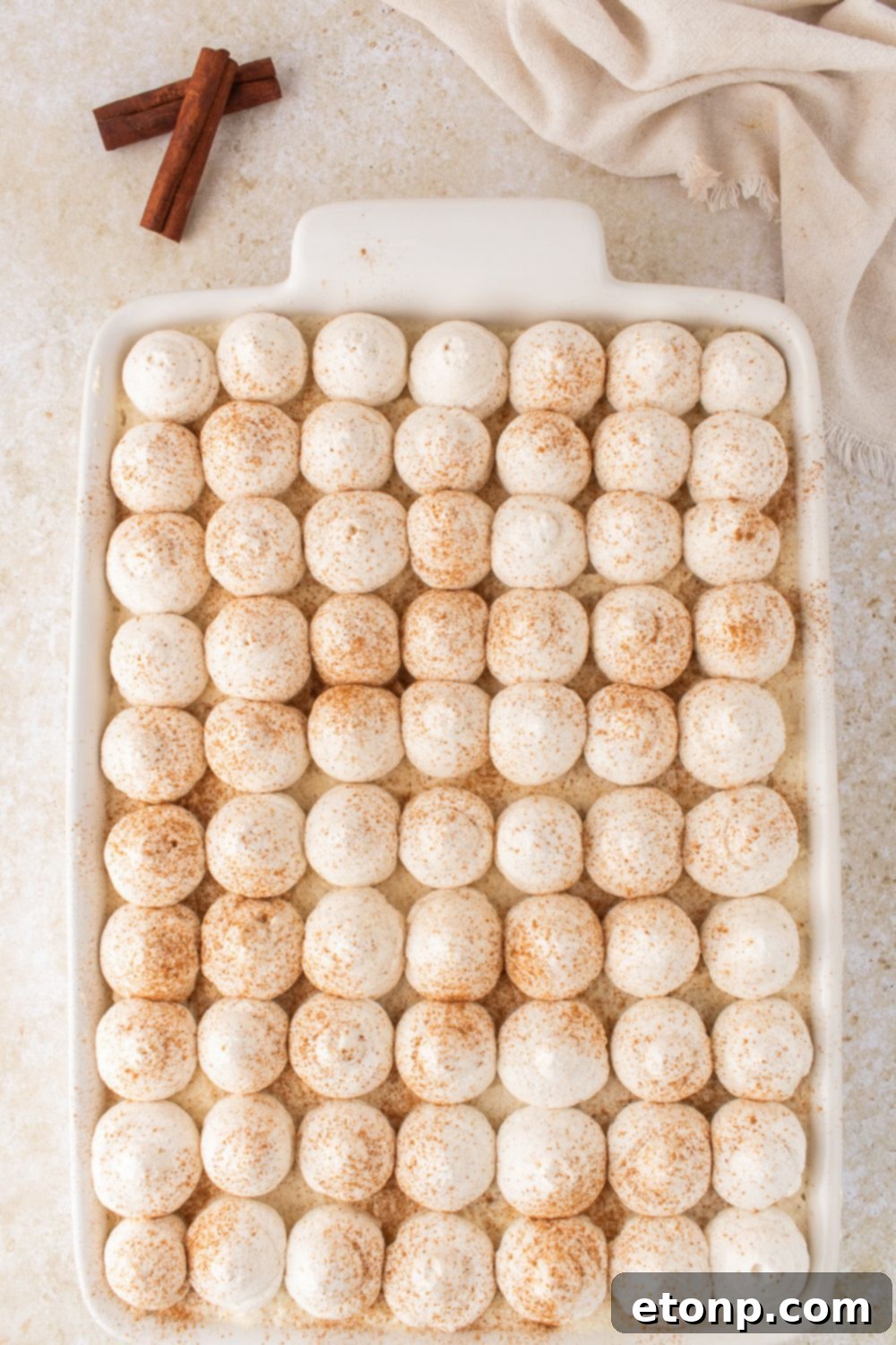 Eggnog Tiramisu: A Festive Holiday Treat 15 Eggnog Tiramisu in a white casserole dish on table with cinnamon sticks to the side
