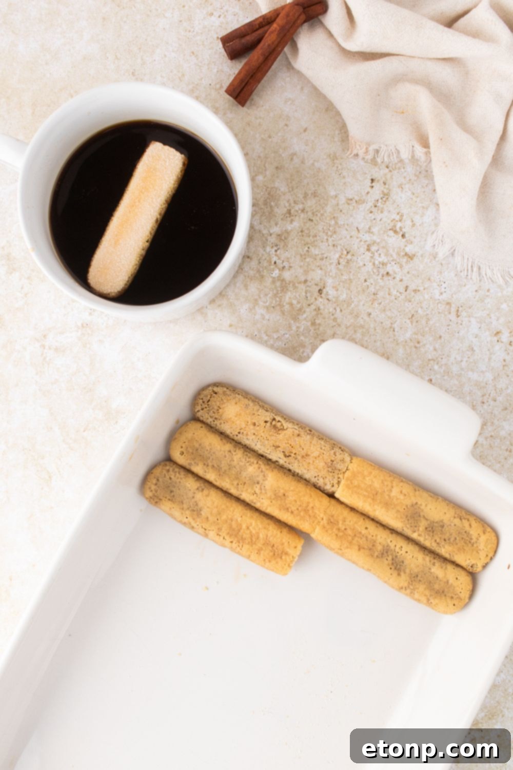 Eggnog Tiramisu: A Festive Holiday Treat 9 A bowl of coffee to side for dipping ladyfinger cookies with a casserole dish with a layer of cookies being placed in it