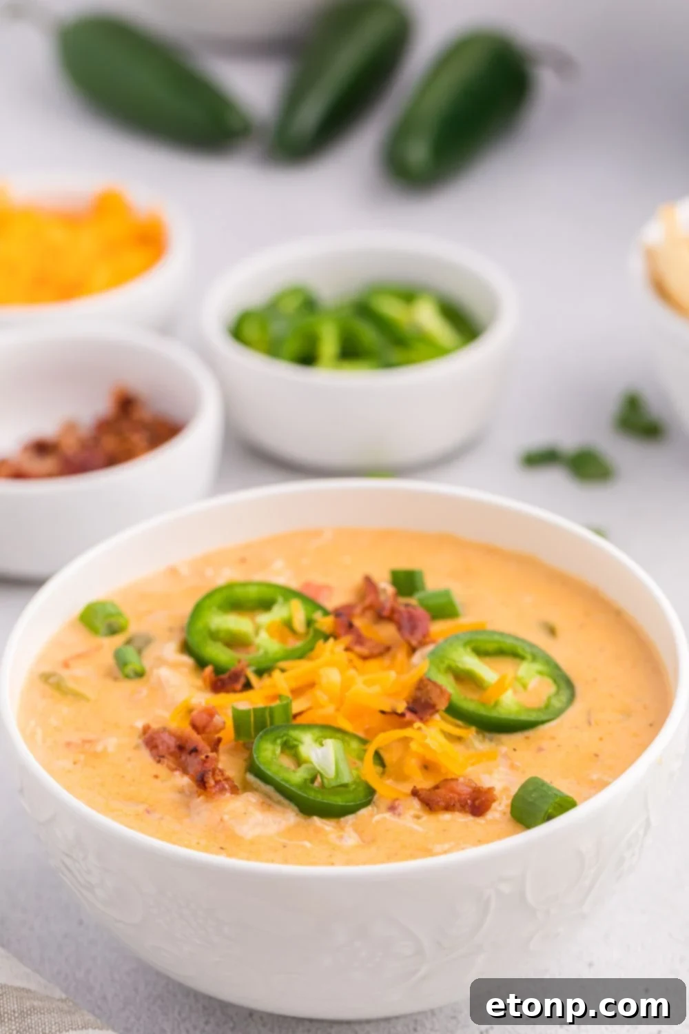 Jalapeño popper crack chicken soup in a white bowl with more of the ingredients around the table and whole jalapeños behind it on table