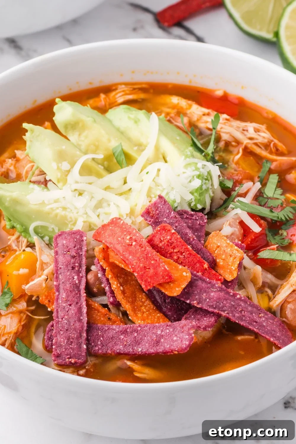 close up look of white bowl full of Southwestern Chicken Soup topped with tortilla strips, shredded cheese and sliced avocado 