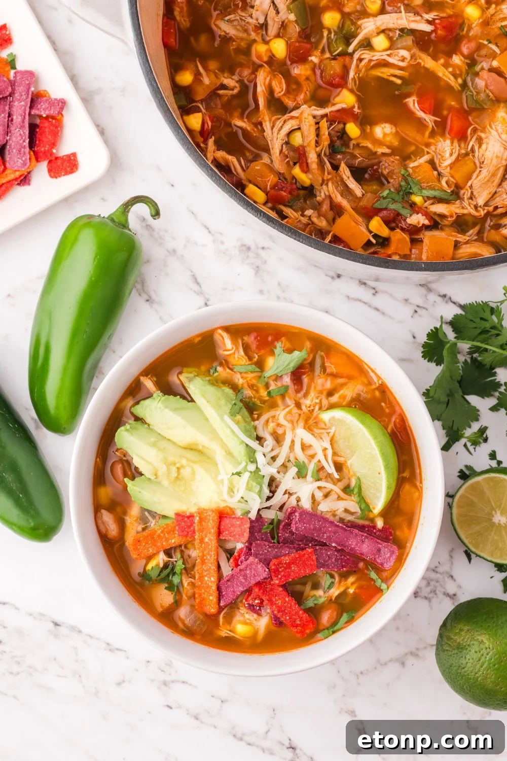 Southwestern Chicken Soup in a white bowl on the table and a pot farther back on the table topped with avocado, chips, cheese, and whole jalapenos on table 
