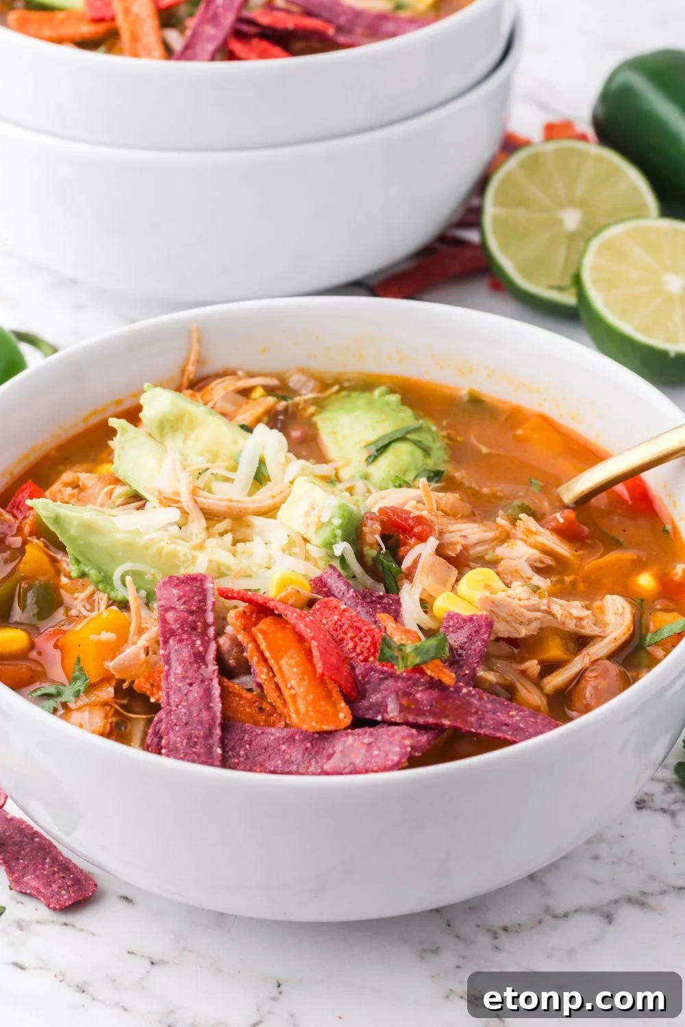 white bowl on table full of Southwestern Chicken Soup topped with avocado, shredded cheese, tortilla chips and more with more bowls stacked in the back of table with limes on the table 