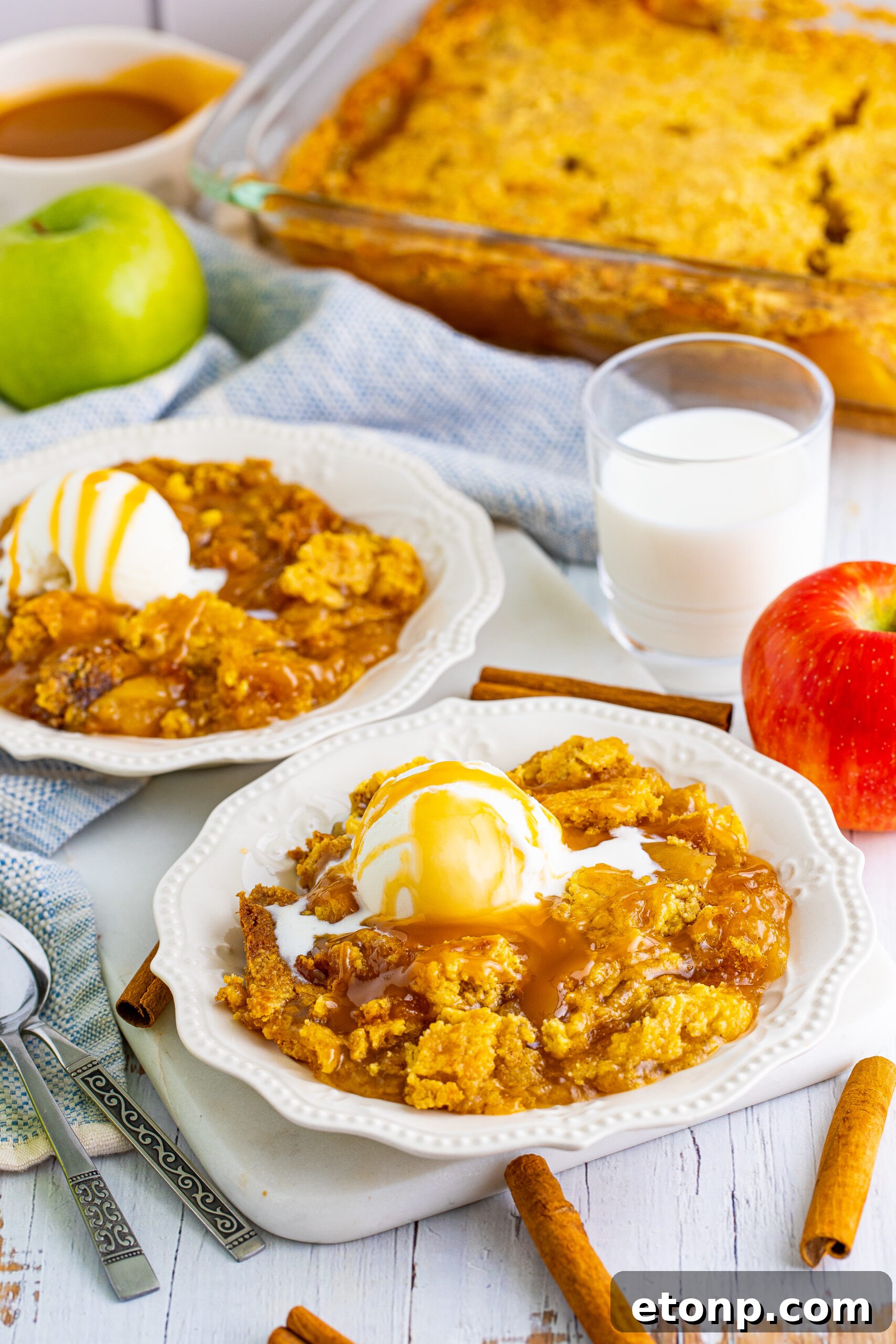 Close up shot of caramel apple dump cake on two white plates with cinnamon sticks and apples in the background.